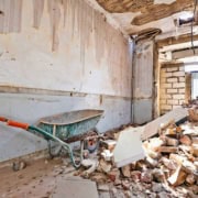 Indendørsbillede af et rum under renovering med en trillebør, murbrokker, afskallet tapet og ødelagt loft